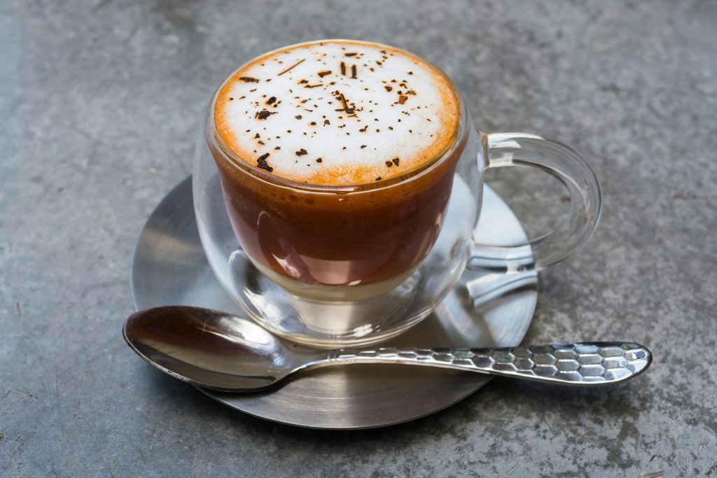 Rich aroma cappuccino in a clear glass mug with frothy foam and sprinkled chocolate on a metallic saucer.